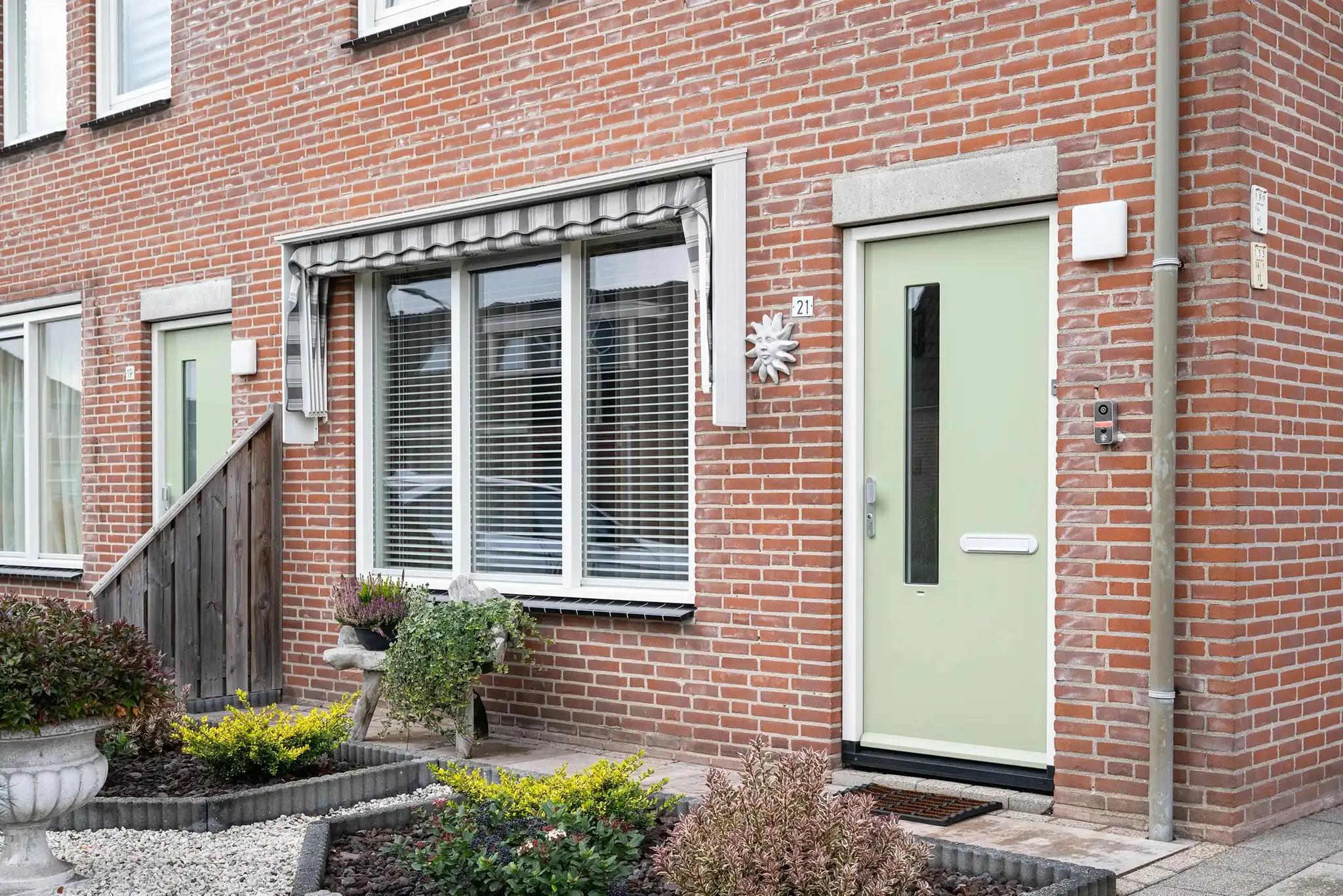 Zachtgroene voordeur naast een raam met markies en een verzorgde voortuin
